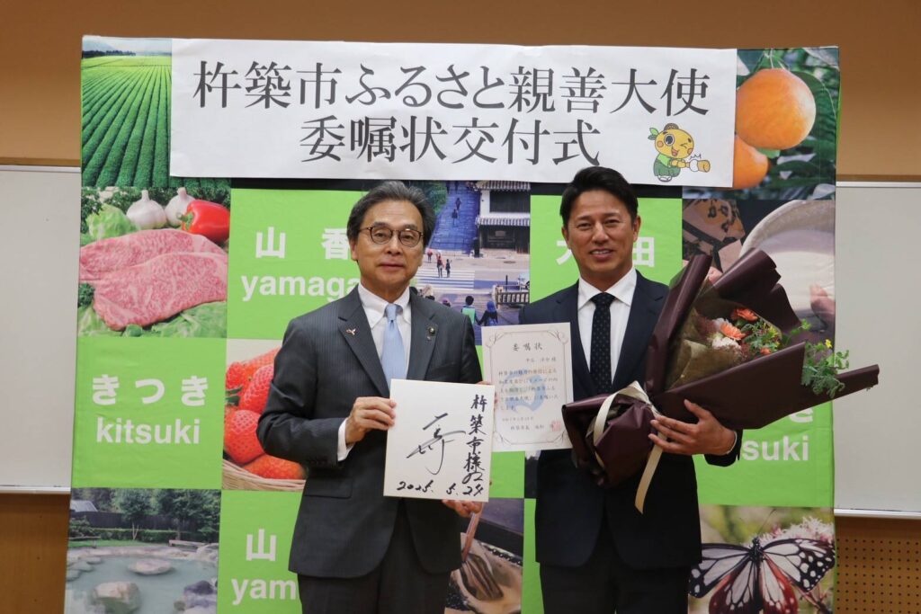 「杵築市ふるさと親善大使」委嘱状交付式の様子。左はスーツ姿の杵築市永松市長、右は花束と委嘱状を持つ楽天イーグルス元監督・平石洋介氏。背景には特産品や観光地の写真が並び、地域の魅力が紹介されている。