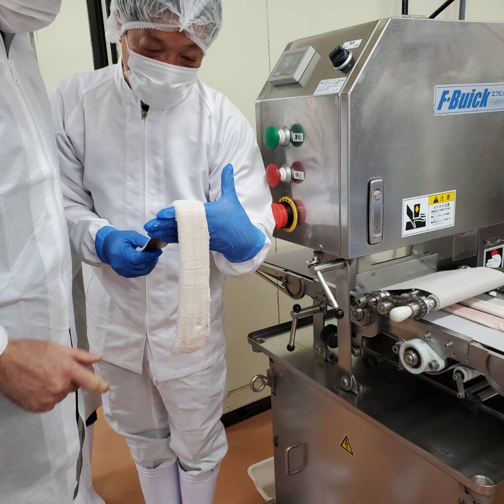 白衣と衛生帽、手袋を着用した作業員が、ハモ（鱧）の加工を行っている様子。右には食品加工機が見え、整形されたハモの身が機械から出ている。