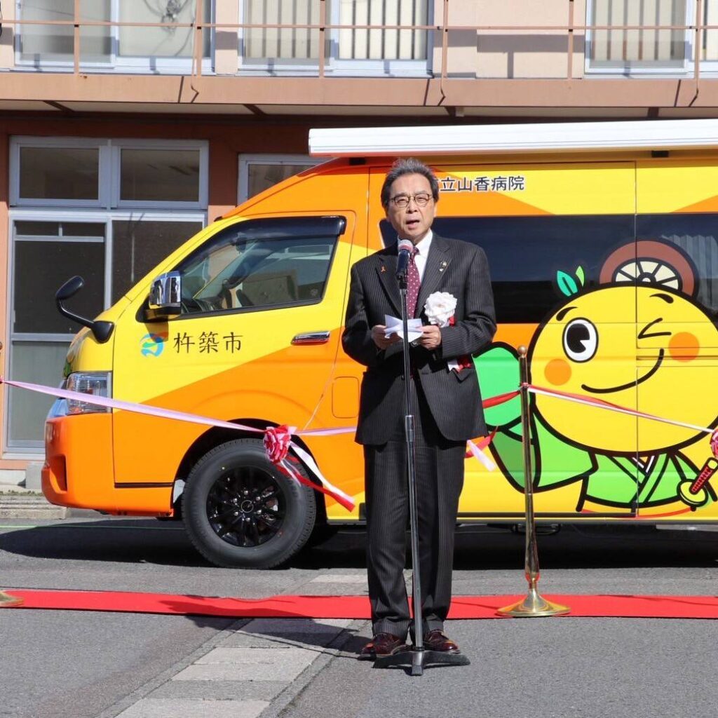 医療MaaS車両の前で挨拶をする永松市長。カラフルな車両には杵築市のロゴと、マスコットキャラクターが描かれており、赤いリボンカット用テープが張られたセレモニーの様子。